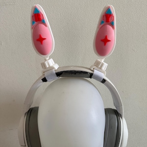 May include: White headphones with gray earcups and attached bunny ears. The bunny ears are white with pink centers and red and blue accents. A red star is on each pink center. The headphones are on a white mannequin head.