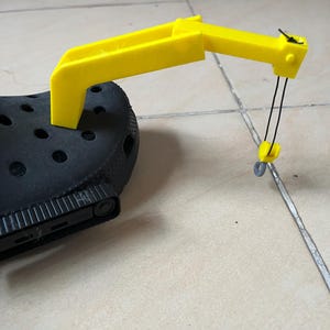 May include: A yellow 3D-printed crane attachment for a black clog shoe. The crane has a hook suspended by black strings. The crane is attached to the top of the shoe, with the crane arm extending outwards.