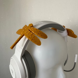 May include: White headphones with a grey earcup and a microphone. The headphones have a yellow dragonfly-shaped accessory attached to the headband.