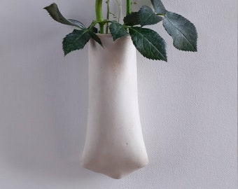May include: A white ceramic vase with a unique shape hangs on a wall with two white roses and green leaves.