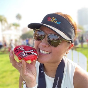 May include: A woman wearing a black visor with the text "Sun Run Track Club" embroidered on it, sunglasses, a white tank top, and a black lulu lemon lanyard with a gold rabbit charm. She is holding a red and white donut with black sprinkles and chocolate drizzle.