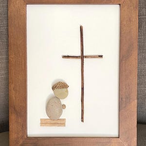May include: Framed artwork featuring a cross and a figure made of natural materials. The cross is constructed from twigs, and the figure is made of pebbles and an acorn. The frame is made of brown wood, and the background is white.