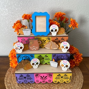 May include: A colorful Day of the Dead altar with multiple tiers, adorned with marigold flowers, sugar skull figurines, and miniature food items. A blue picture frame sits at the top. The altar is placed on a woven mat.
