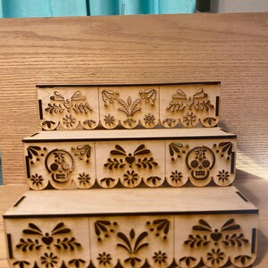 May include: Three-tiered wooden display shelves with laser-cut decorative designs. The designs include floral patterns, hearts, and skull motifs. The shelves are made of light-colored wood and are likely for displaying small items or collectibles.