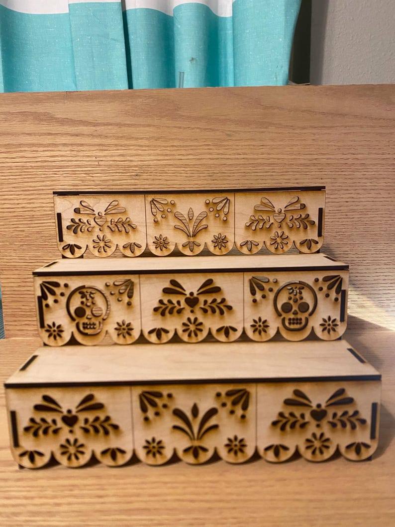 May include: Three-tiered wooden display shelves with laser-cut decorative designs. The designs include floral patterns, hearts, and skull motifs. The shelves are made of light-colored wood and are likely for displaying small items or collectibles.