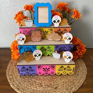 May include: A multi-tiered, colorful Day of the Dead altar display. The wooden structure features vibrant paint and decorative cutouts. Adorned with marigold flowers, miniature skulls, food replicas, and a blue picture frame, it sits on a woven mat.