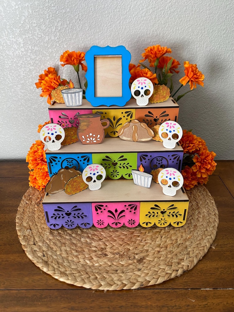 May include: A multi-tiered, colorful Day of the Dead altar display. The wooden structure features vibrant paint and decorative cutouts. Adorned with marigold flowers, miniature skulls, food replicas, and a blue picture frame, it sits on a woven mat.