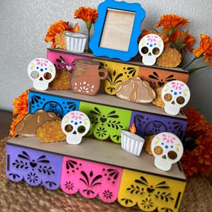 May include: A multi-tiered wooden display featuring Day of the Dead themed decorations. The display includes colorful panels, sugar skull cutouts, a blue picture frame, and orange marigold flowers. The overall design is festive and decorative.