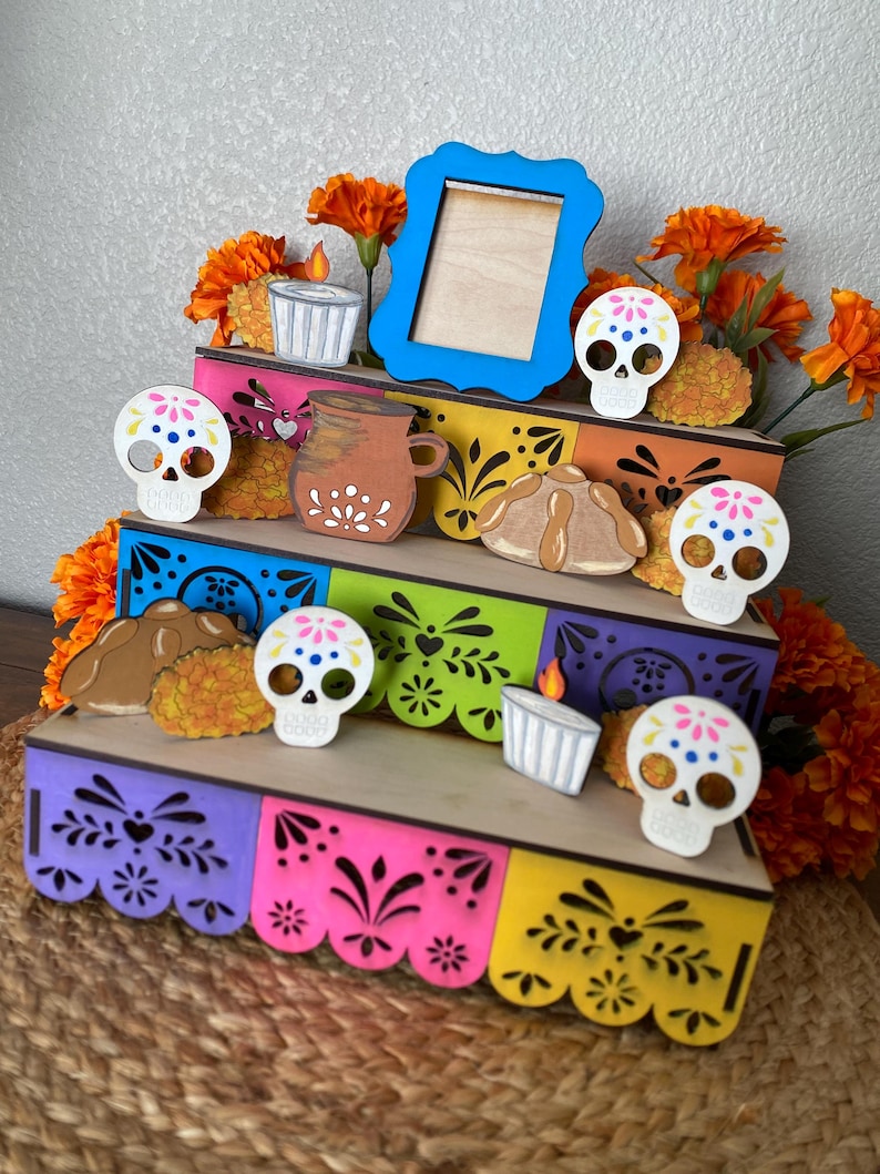 May include: A multi-tiered wooden display featuring Day of the Dead themed decorations. The display includes colorful panels, sugar skull cutouts, a blue picture frame, and orange marigold flowers. The overall design is festive and decorative.