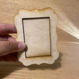 May include: A small, unfinished wooden picture frame with a decorative, scalloped edge. The frame has a rectangular opening and is held by a hand. The wood has a natural, light brown color.