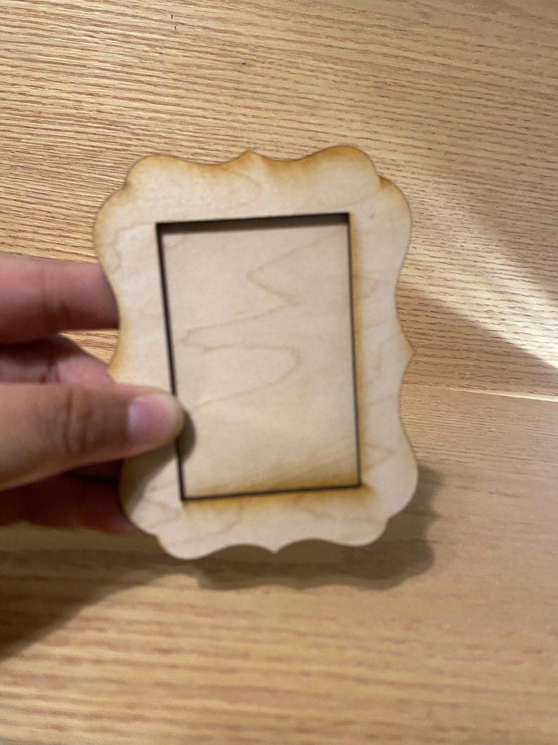 May include: A small, unfinished wooden picture frame with a decorative, scalloped edge. The frame has a rectangular opening and is held by a hand. The wood has a natural, light brown color.