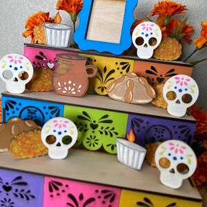 May include: A Day of the Dead themed wooden display featuring a blue picture frame, sugar skull cutouts, and decorative elements. The display includes a tiered structure with colorful cut-out designs and orange flowers.