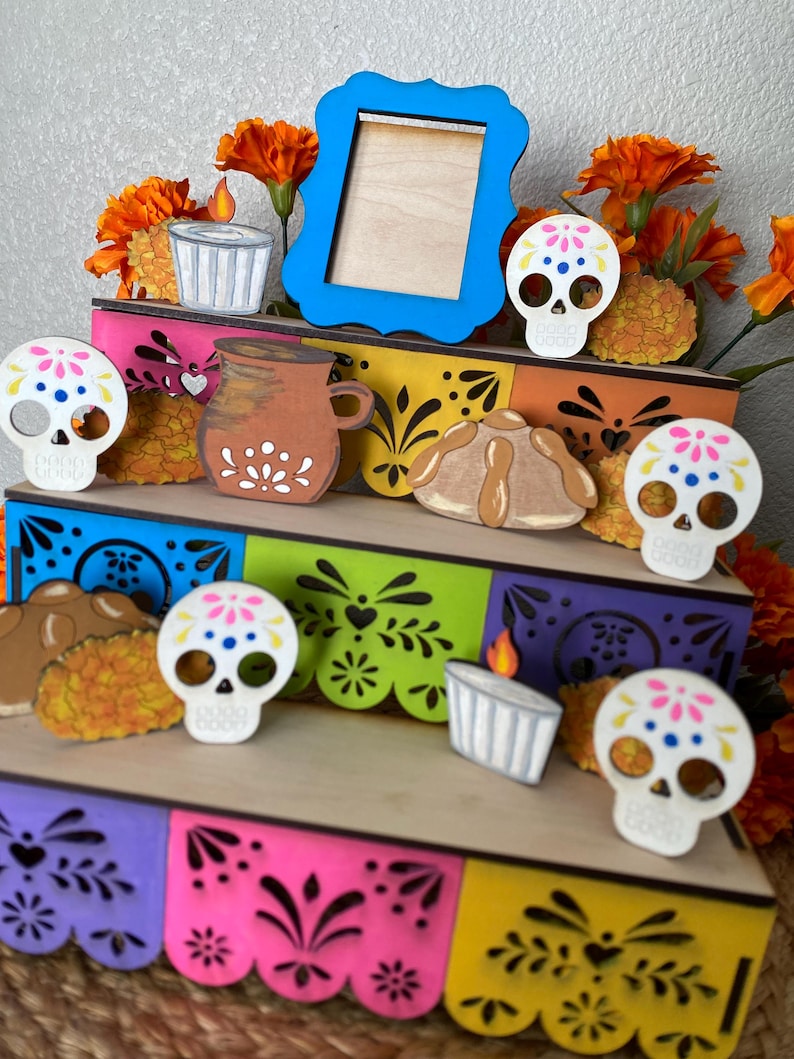 May include: A Day of the Dead themed wooden display featuring a blue picture frame, sugar skull cutouts, and decorative elements. The display includes a tiered structure with colorful cut-out designs and orange flowers.