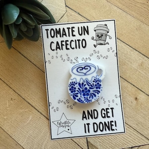 May include: A white and blue ceramic coffee cup pin with floral designs and a heart-shaped coffee art design. The pin is on a card with the text "TOMATE UN CAFECITO AND GET IT DONE!" and coffee bean illustrations.