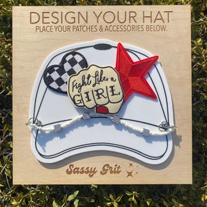 May include: A white baseball cap with a design your own hat theme. The cap has a red star, a black and white checkered heart, a white beaded chain with black and white checkered beads, and a patch that says "Fight Like a Girl". The text "Sassy Grit" is engraved on the wooden background.