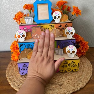 May include: A Day of the Dead altar with three colorful wooden tiers. The altar is decorated with orange flowers, sugar skulls, candles, and bread. A blue picture frame sits on the top tier. The altar is on a woven mat.