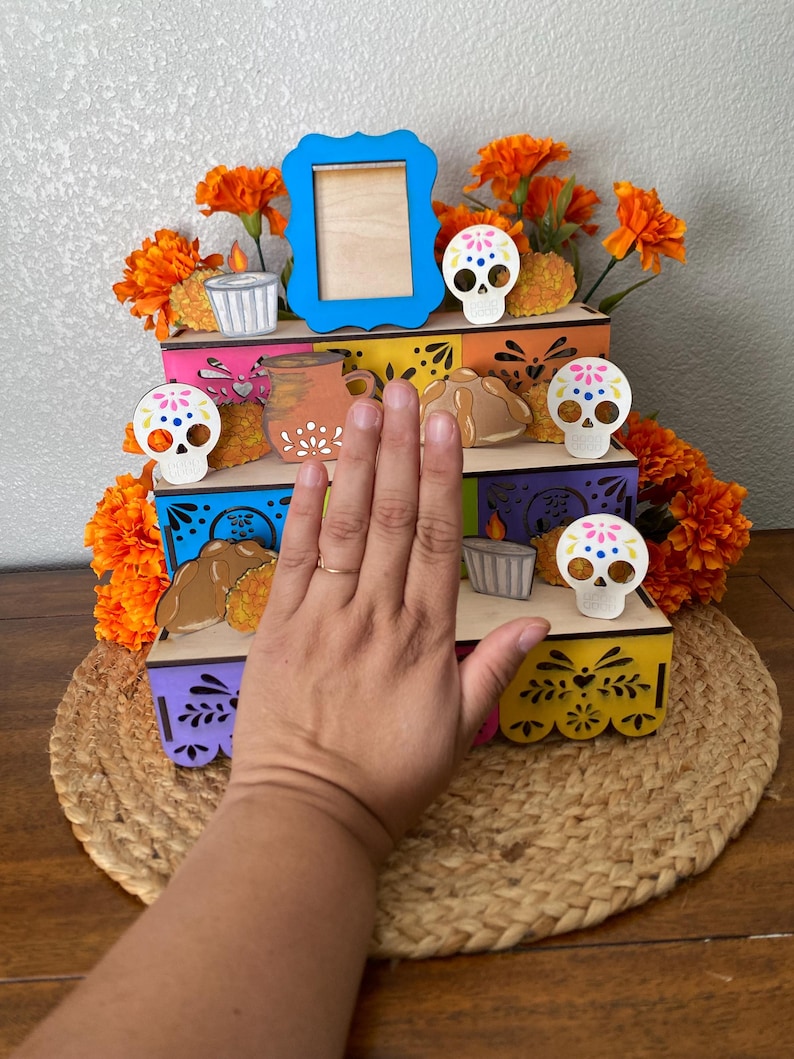 May include: A Day of the Dead altar with three colorful wooden tiers. The altar is decorated with orange flowers, sugar skulls, candles, and bread. A blue picture frame sits on the top tier. The altar is on a woven mat.