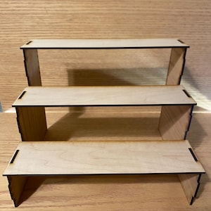 May include: Three-tiered wooden display stand. Each level is a rectangular platform supported by two side pieces. The wood has a light, natural color with dark edges. The stand is designed to showcase items.