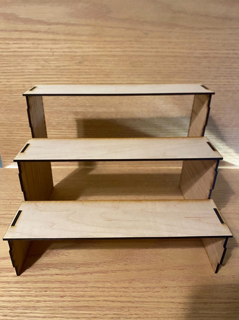 May include: Three-tiered wooden display stand. Each level is a rectangular platform supported by two side pieces. The wood has a light, natural color with dark edges. The stand is designed to showcase items.