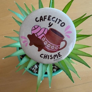 May include: A circular pin-back button with a light beige background featuring a brown mug, a pink concha bread, and a dark brown cookie. The text reads "CAFECITO y CHISME" in dark brown letters, accented by small pink decorative splashes.