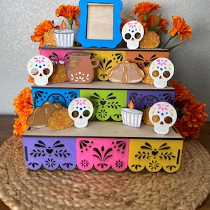 May include: A multi-tiered, colorful Day of the Dead display. The display features a blue picture frame, wooden skull decorations, candles, and marigold flowers. The display is on a woven mat.