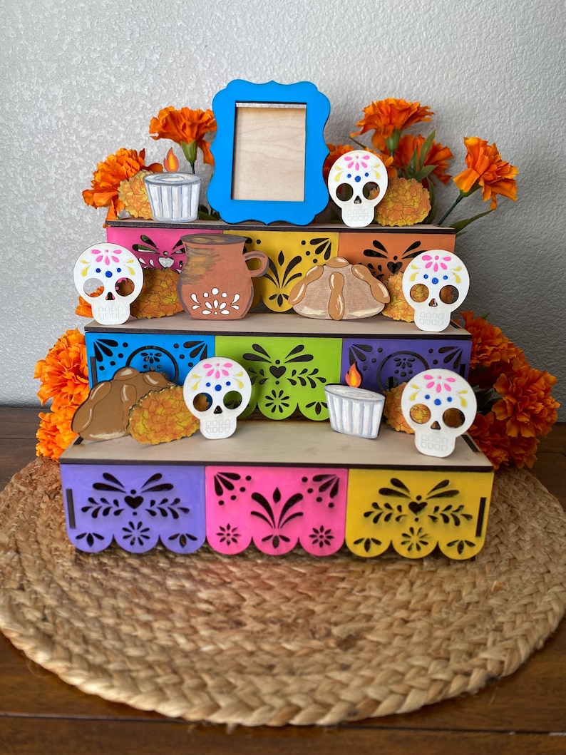 May include: A multi-tiered, colorful Day of the Dead display. The display features a blue picture frame, wooden skull decorations, candles, and marigold flowers. The display is on a woven mat.