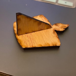 May include: A wooden phone stand with a natural wood grain finish. The stand features a triangular support and a decorative wing-shaped element. The wood has a dark, burnt edge, adding contrast to the light brown color.