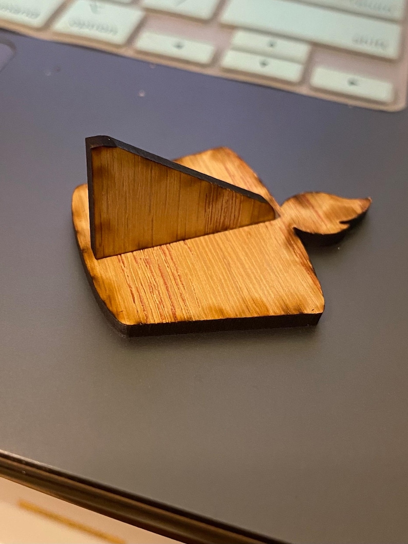 May include: A wooden phone stand with a natural wood grain finish. The stand features a triangular support and a decorative wing-shaped element. The wood has a dark, burnt edge, adding contrast to the light brown color.