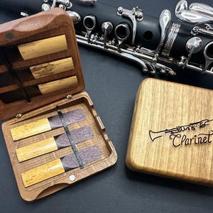 Puede incluir: Una caja de madera con tapa abatible, que contiene un juego de cañas de clarinete. La caja está abierta, revelando las cañas en el interior. La caja está grabada con la palabra "Clarinete" y una simple ilustración de un clarinete.