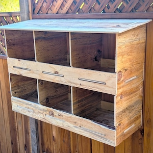 May include: A wooden chicken coop with a slanted roof and multiple compartments. The coop is constructed from light-colored wood and features a natural wood grain. It is mounted on a wooden fence.