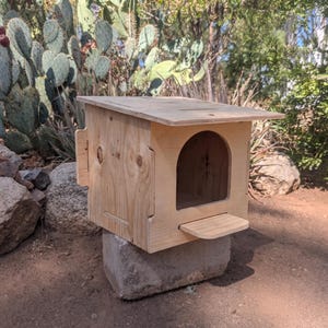 May include: A light-colored wooden animal shelter featuring a flat, overhanging roof, a single arched entrance, and a small rectangular landing platform extending from the front. The structure is constructed from plywood panels with visible wood grain.