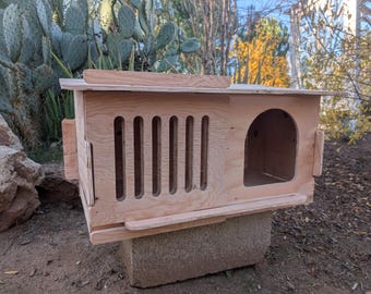 Puede incluir: Una pajarera de madera con forma rectangular y techo inclinado. La pajarera tiene una entrada redondeada y ranuras verticales en un lado. Está construida con madera de color claro y se asienta sobre un bloque de hormigón.