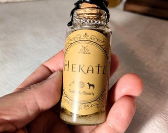 Könnte beinhalten: Kleine Glasflasche mit Korken und schwarzem Wachssiegel. Das Etikett trägt die Aufschrift "Hekate" mit einem Hund und anderen Symbolen. Die Flasche enthält eine hellfarbene, körnige Substanz.
