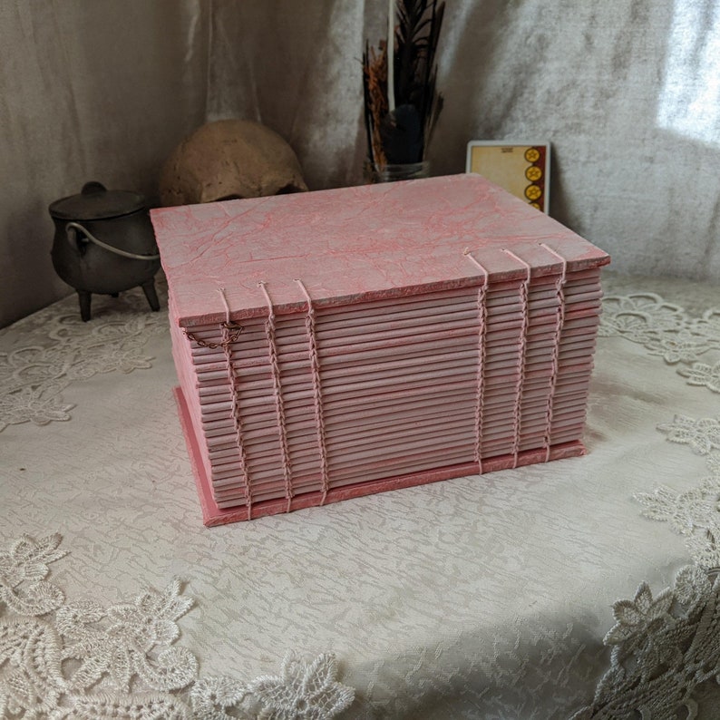 May include: A handmade pink journal with a textured cover and stitched binding. The pages are visible along the edges, and the journal is rectangular in shape. The cover has a marbled appearance, and the binding is a lighter pink.