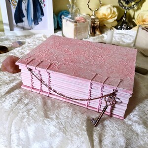 May include: A handmade pink journal with a textured cover and hand-stitched binding. A decorative chain with a key charm secures the journal. The journal is approximately 8 inches long and 6 inches wide.