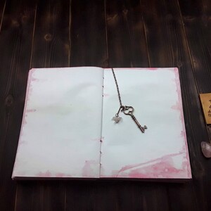 May include: Open journal with pink watercolor edges, a vintage key bookmark, and a small wooden tag. The key is bronze-colored and has a small crystal charm. The journal is open on a dark wood surface.