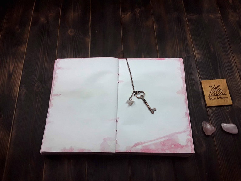 May include: Open journal with pink watercolor edges, a vintage key bookmark, and a small wooden tag. The key is bronze-colored and has a small crystal charm. The journal is open on a dark wood surface.