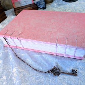 May include: A handmade pink journal with a textured cover and hand-stitched binding. A decorative bronze key charm hangs from the journal's spine. The journal's pages are visible, and the overall aesthetic is vintage and artisanal.