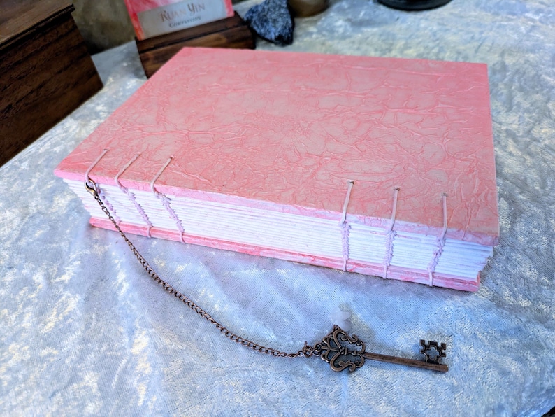 May include: A handmade pink journal with a textured cover and hand-stitched binding. A decorative bronze key charm hangs from the journal's spine. The journal's pages are visible, and the overall aesthetic is vintage and artisanal.