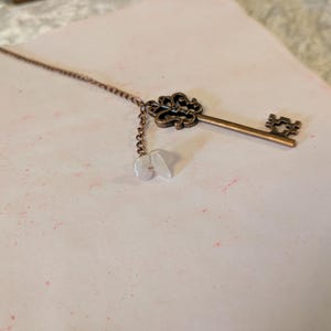 May include: A bronze-colored key pendant with a decorative crown-like top and a chain with three small, white, irregular-shaped beads. The key lies on a light pink surface with subtle red speckles.