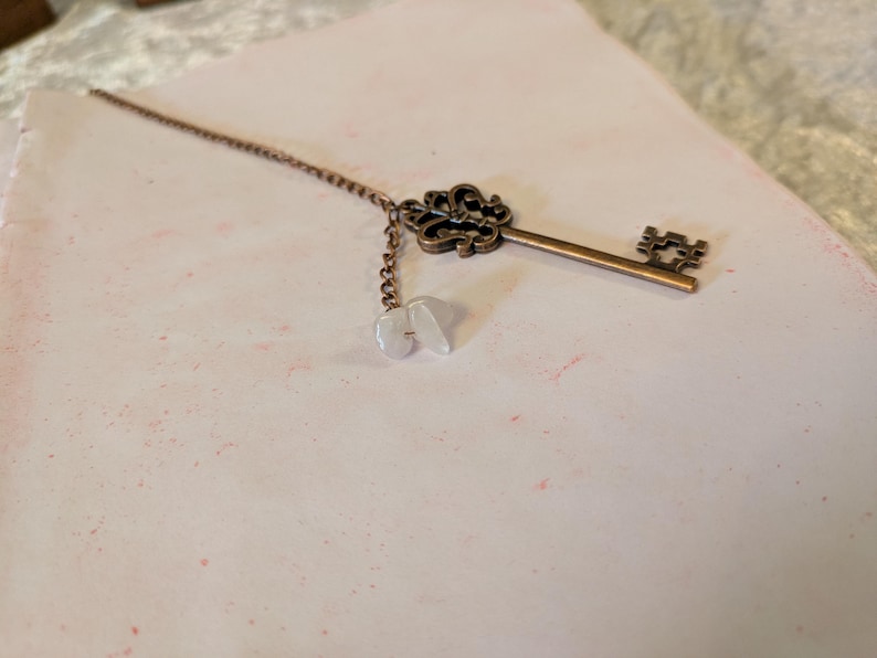 May include: A bronze-colored key pendant with a decorative crown-like top and a chain with three small, white, irregular-shaped beads. The key lies on a light pink surface with subtle red speckles.