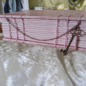 May include: A pink, handmade book-shaped box with a decorative key pendant. The box is constructed from layered, textured paper and bound with pink thread. A bronze-colored key hangs from a chain attached to the box, adding a vintage aesthetic.