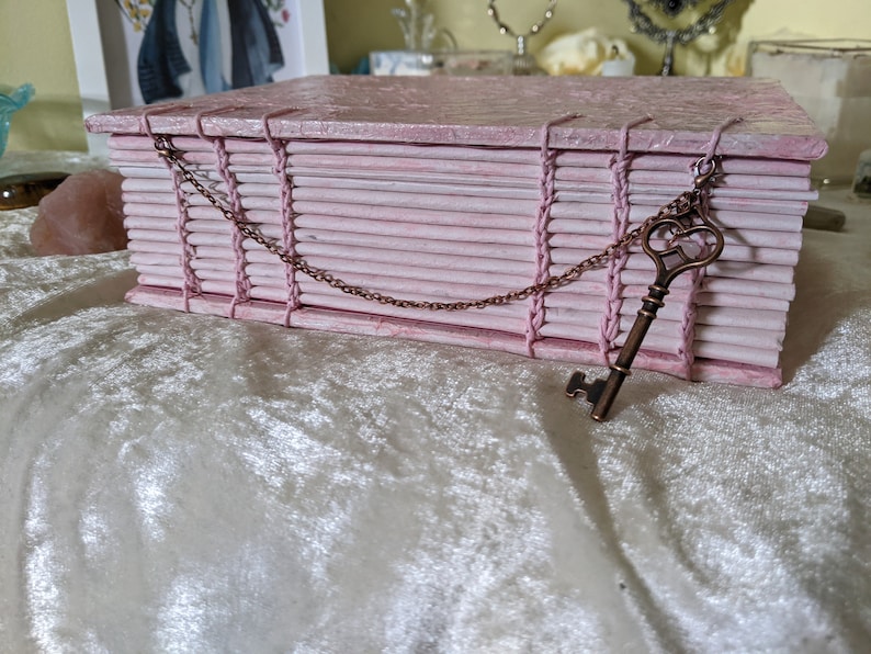 May include: A pink, handmade book-shaped box with a decorative key pendant. The box is constructed from layered, textured paper and bound with pink thread. A bronze-colored key hangs from a chain attached to the box, adding a vintage aesthetic.