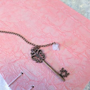 May include: A vintage-style key pendant with a decorative head and a small gemstone charm hangs on a chain. The key rests on a textured pink surface, possibly a journal or notebook cover. The key is made of a bronze-colored metal.