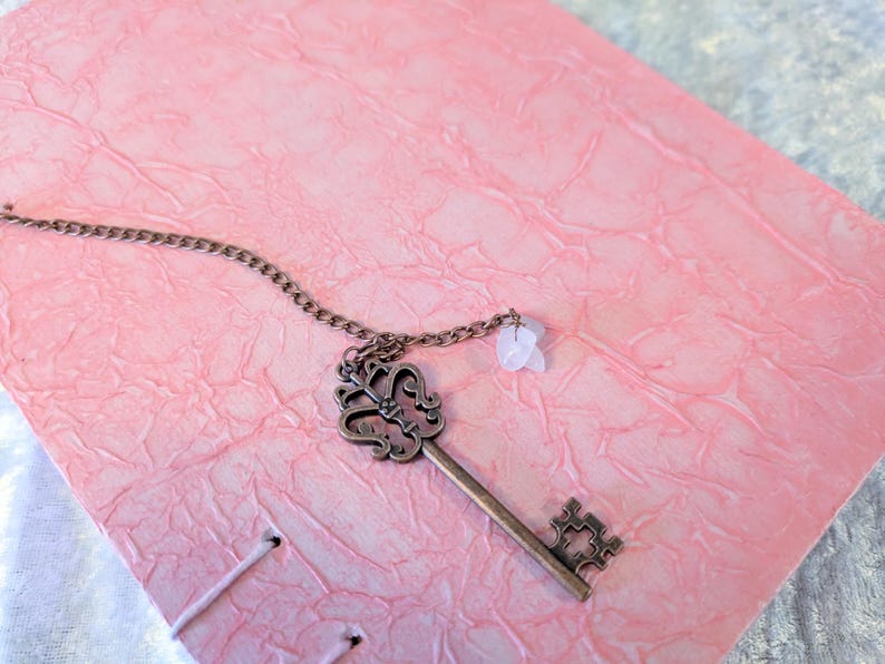 May include: A vintage-style key pendant with a decorative head and a small gemstone charm hangs on a chain. The key rests on a textured pink surface, possibly a journal or notebook cover. The key is made of a bronze-colored metal.
