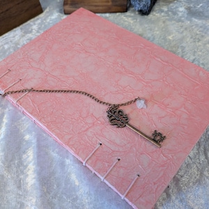 May include: A pink journal with a textured cover and stitched binding. A decorative bronze key pendant with a small white charm rests on the cover. The journal is closed and lies on a light-colored surface.