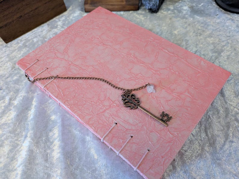 May include: A pink journal with a textured cover and stitched binding. A decorative bronze key pendant with a small white charm rests on the cover. The journal is closed and lies on a light-colored surface.