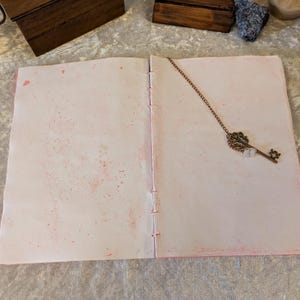 May include: Open journal with pink-splattered pages and a decorative key-shaped bookmark. The journal's pages are stitched together with pink thread. The key bookmark is made of metal and has an ornate design.