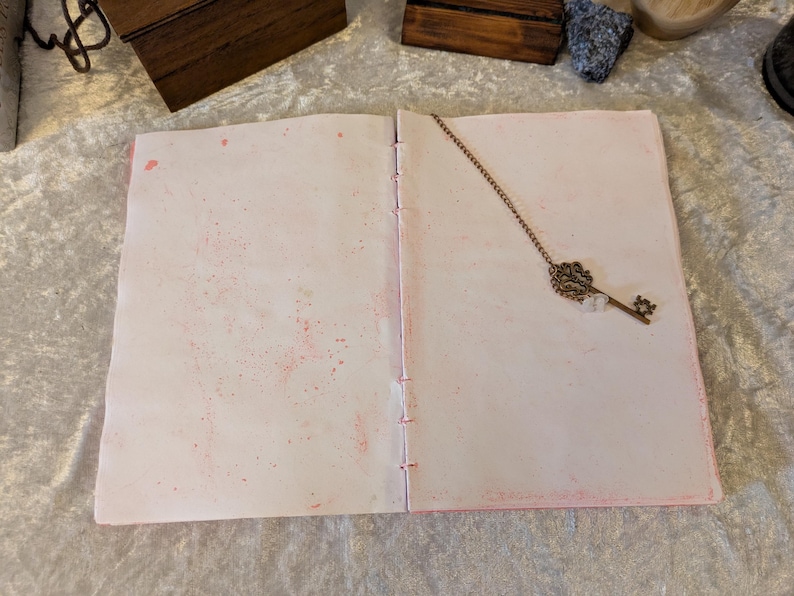 May include: Open journal with pink-splattered pages and a decorative key-shaped bookmark. The journal's pages are stitched together with pink thread. The key bookmark is made of metal and has an ornate design.