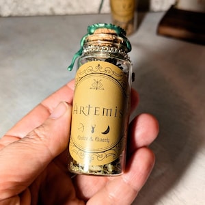 Peut inclure: Une petite bouteille en verre avec un bouchon en liège et un sceau de cire vert. La bouteille est remplie d'herbes et de cristaux. L'étiquette sur la bouteille indique "Artemis" avec un arc et une flèche, un cerf et une lune croissante. L'étiquette indique également "Quire & Quartz".